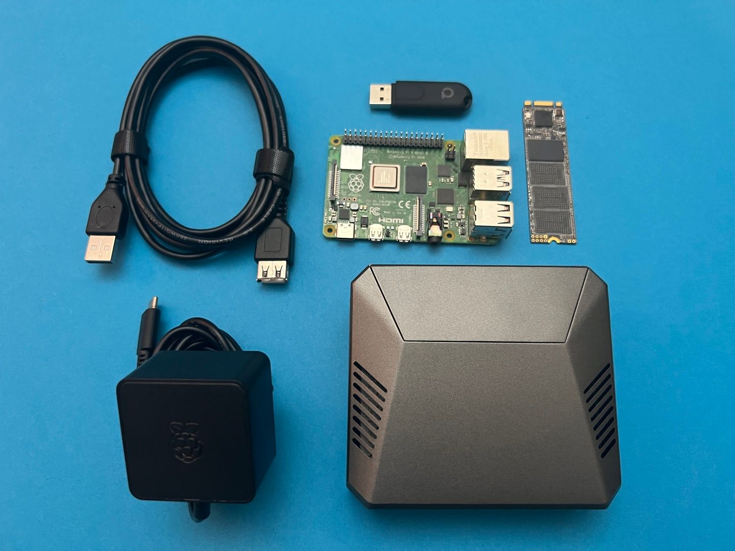 A Raspberry Pi 4 Model B next to its USB-C power supply, an USB extension cable, a USB zigbee dongle, and an M.2 SSD adapter on a blue background